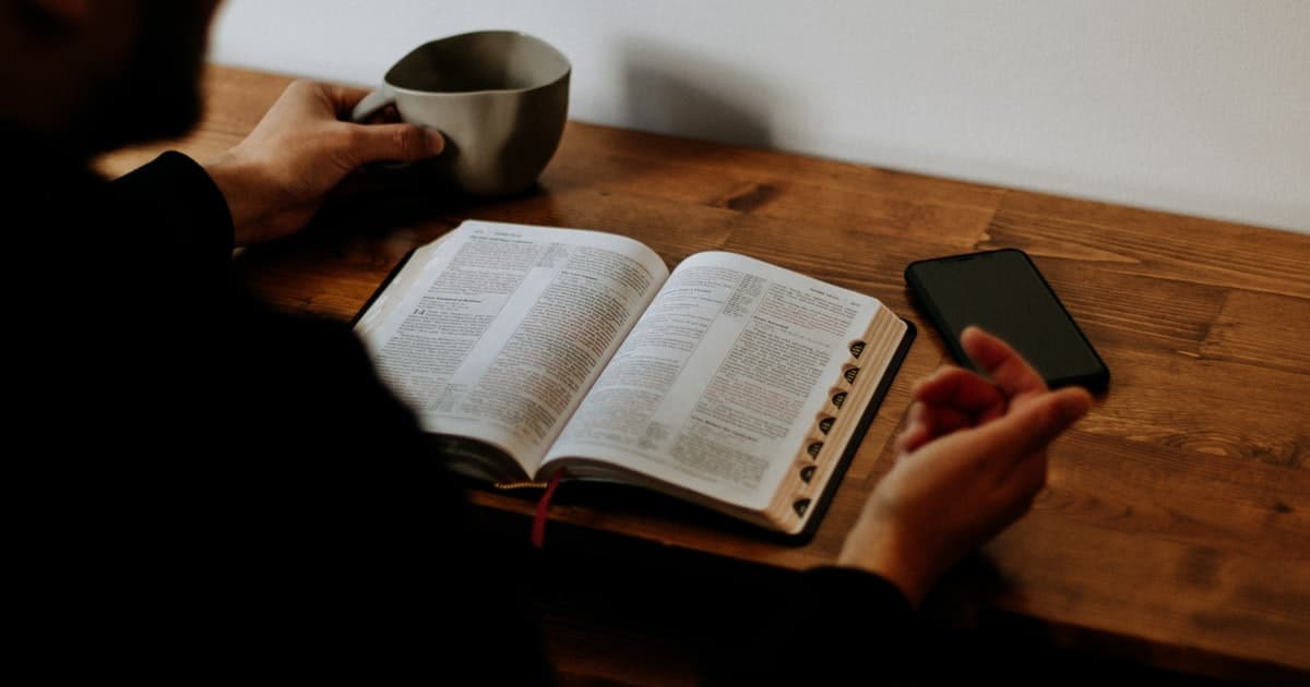 person reading Bible with coffee and phone on table