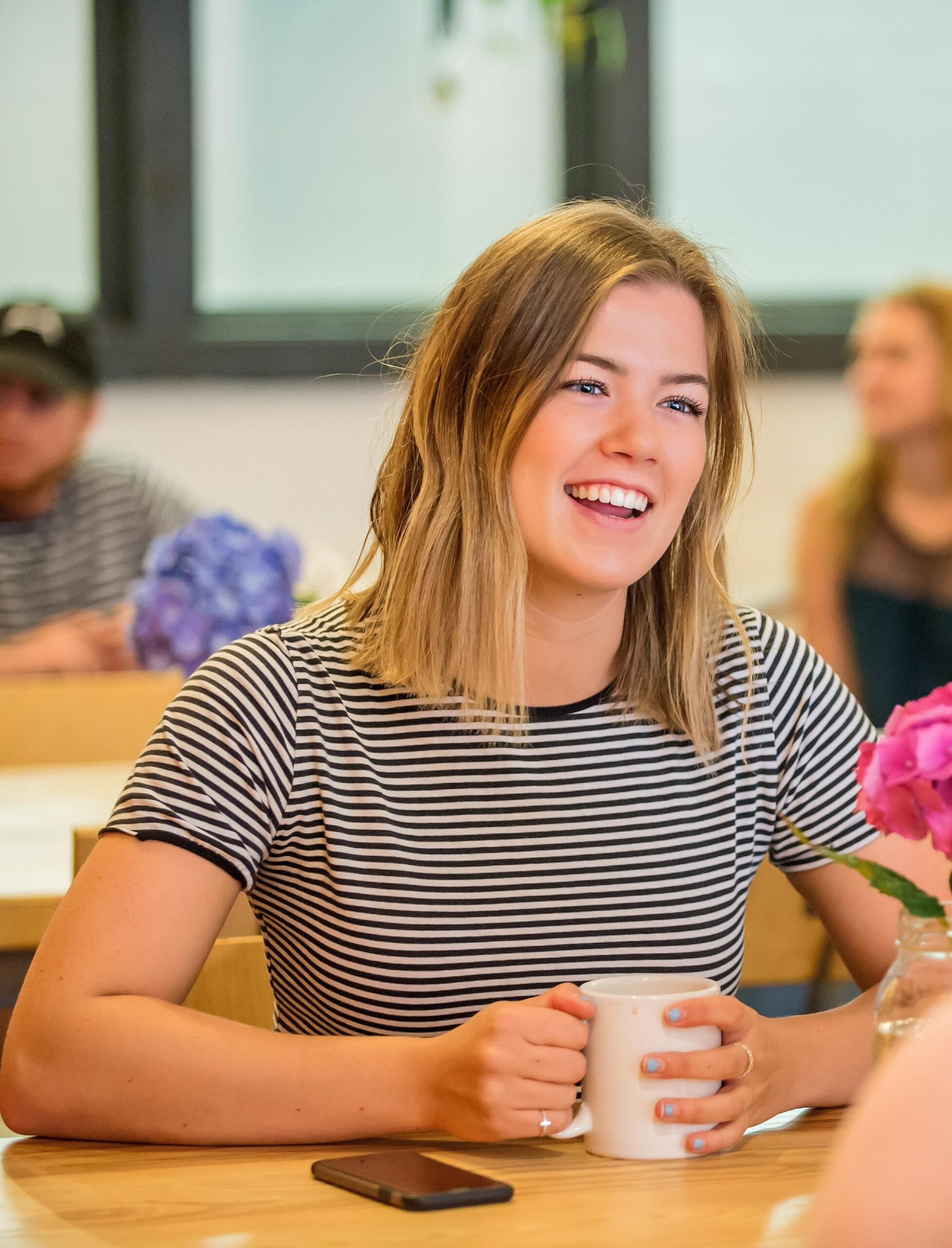 smiling female student at a table in commons area with phone and cup of coffee