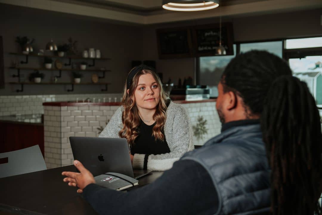 Two students sitting at a table in front of a laptop talking together